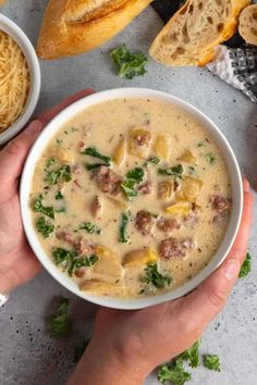 two hands holding a bowl of soup with cheese and spinach on the table next to bread