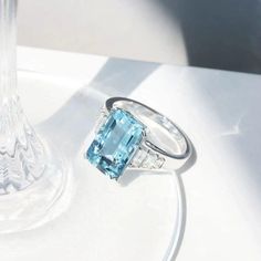 a ring with an aqua blue topaz sits next to a clear glass vase on a white surface