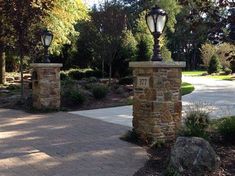 two street lamps on top of stone pillars in front of trees and bushes, along with a brick walkway