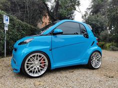 a blue smart car parked on gravel next to trees