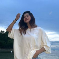 a woman standing next to the water with her hands in her hair and wearing a white shirt