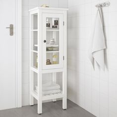 a white bathroom cabinet with glass doors and shelves on the wall next to a towel rack