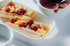 two banana split desserts with fruit and nuts in them on a white plate next to a cup of coffee