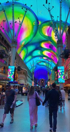 people walking through an indoor shopping mall with colorful lights on the ceiling and overhead lighting