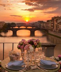 the table is set with flowers and wine glasses on it in front of an old bridge