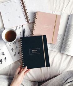 a person holding a notebook next to two books and a cup of coffee on a bed