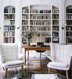 two white chairs sitting in front of a wooden table and bookshelf filled with lots of books