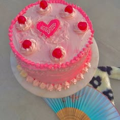 a cake with pink frosting and hearts on top, sitting next to a fan