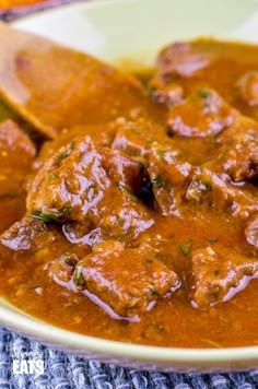 a white bowl filled with meat and sauce on top of a blue table cloth next to a wooden spoon