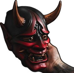 a red demon mask with horns on it's head is held up by a hand