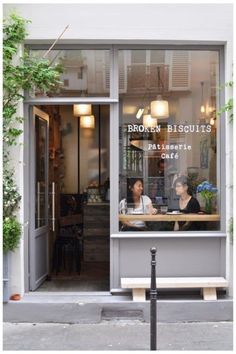 two women sitting at a table in front of a store window with plants growing on the outside