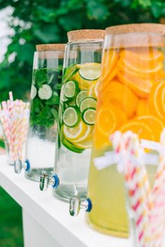 several glasses filled with different types of drinks on a white table in front of trees