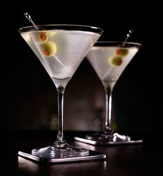 two martini glasses filled with white wine and garnished with olives on a black table