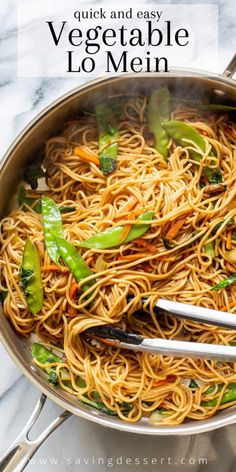 a pan filled with noodles and vegetables on top of a marble countertop next to two tongs