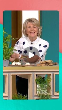 an older woman is smiling while sitting at a table with food and plants in front of her