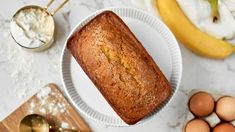 a loaf of bread sitting on top of a white plate next to eggs and bananas