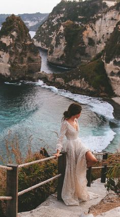 a woman in a white dress standing on the edge of a cliff overlooking the ocean