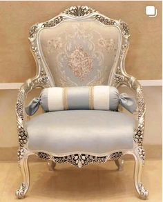 an ornate chair with blue and white pillows on it's back, in front of a beige wall