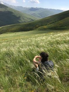 a man sitting in the middle of a lush green field with mountains in the background