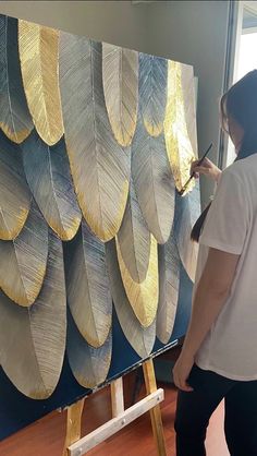 a woman is painting an art piece with gold leaves on the wall behind her, while another person holds a paintbrush in their hand