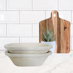 two white bowls sitting on top of a counter next to a cutting board with a plant in it