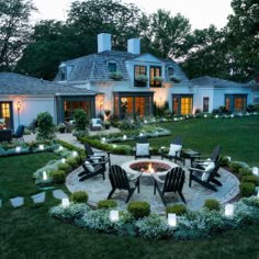 an outdoor fire pit surrounded by lawn furniture and landscaping lights at night in front of a large white house