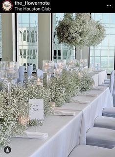 a long table with white flowers and candles in front of large windows at an event