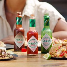 three bottles of tabasco sitting on top of a wooden table next to plates of food