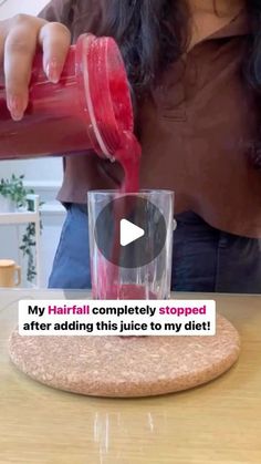 a woman pouring red liquid into a glass on top of a wooden table next to a plant