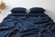 an unmade bed with blue linens and two pillows on it, next to a dried plant