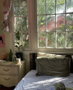 a bed sitting under a window next to a white dresser and table with a potted plant on it