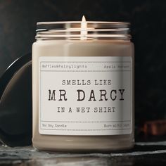 a candle that is sitting on a table next to a black container with the words smells like mr darcy in a wet shirt