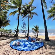 a woman laying on top of a blue and white blanket under palm trees next to the ocean