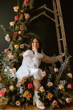 a woman sitting on top of a wooden ladder surrounded by flowers and greenery, posing for the camera