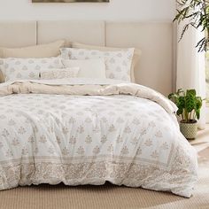 a bed with white comforter and pillows in a room next to a plant on the floor