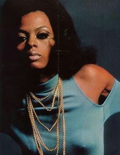 a woman in a blue top with gold chains on her neck and hands behind her back