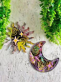 two metal sun and moon wall hangings next to green plants on a white surface