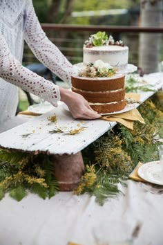 a woman cutting a cake on top of a wooden table with greenery around it