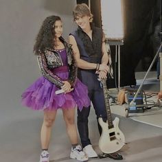 two people standing next to each other in front of a microphone and guitar, with one person wearing a purple dress