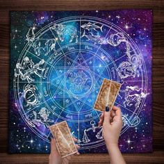 two hands holding tarot cards in front of a painting with zodiac signs on it