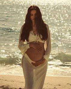 a pregnant woman standing on top of a sandy beach next to the ocean and holding her belly