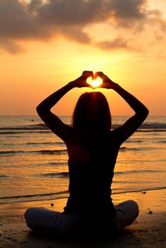 a woman sitting in front of the ocean making a heart shape with her hands at sunset