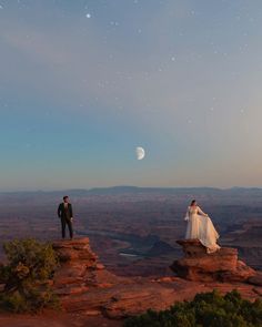 a man and woman standing on top of a cliff looking at the moon in the sky