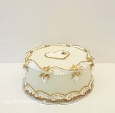 a white cake with gold decorations on top and a heart decoration on the bottom is sitting on a table