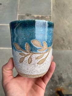 a hand holding a blue and white cup with gold leaves on the outside, in front of a tile floor