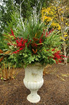 a white vase filled with red and green flowers on top of a dirt ground next to trees