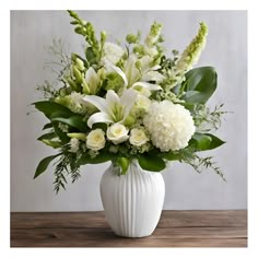 a white vase filled with lots of flowers on top of a wooden table next to a wall