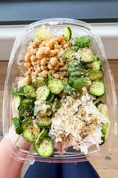 a person holding up a plastic container filled with cucumber and chickpeas