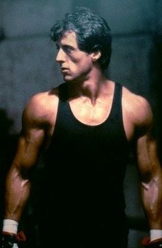 a man in black tank top and gloves standing next to a wall with his hands on his hips