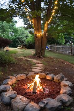 Backyard campfire surrounded by stones, tree wrapped in string lights, and a picnic table in the background along a stone path. Rustic Rock Fire Pit, Whimsical Fire Pit, Natural Fire Pit Ideas, Outdoor Fire Pit Ideas Seating Areas, Sand Fire Pit Area, Simple Fire Pit Ideas, Home Made Fire Pit, Simple Outdoor Fire Pit